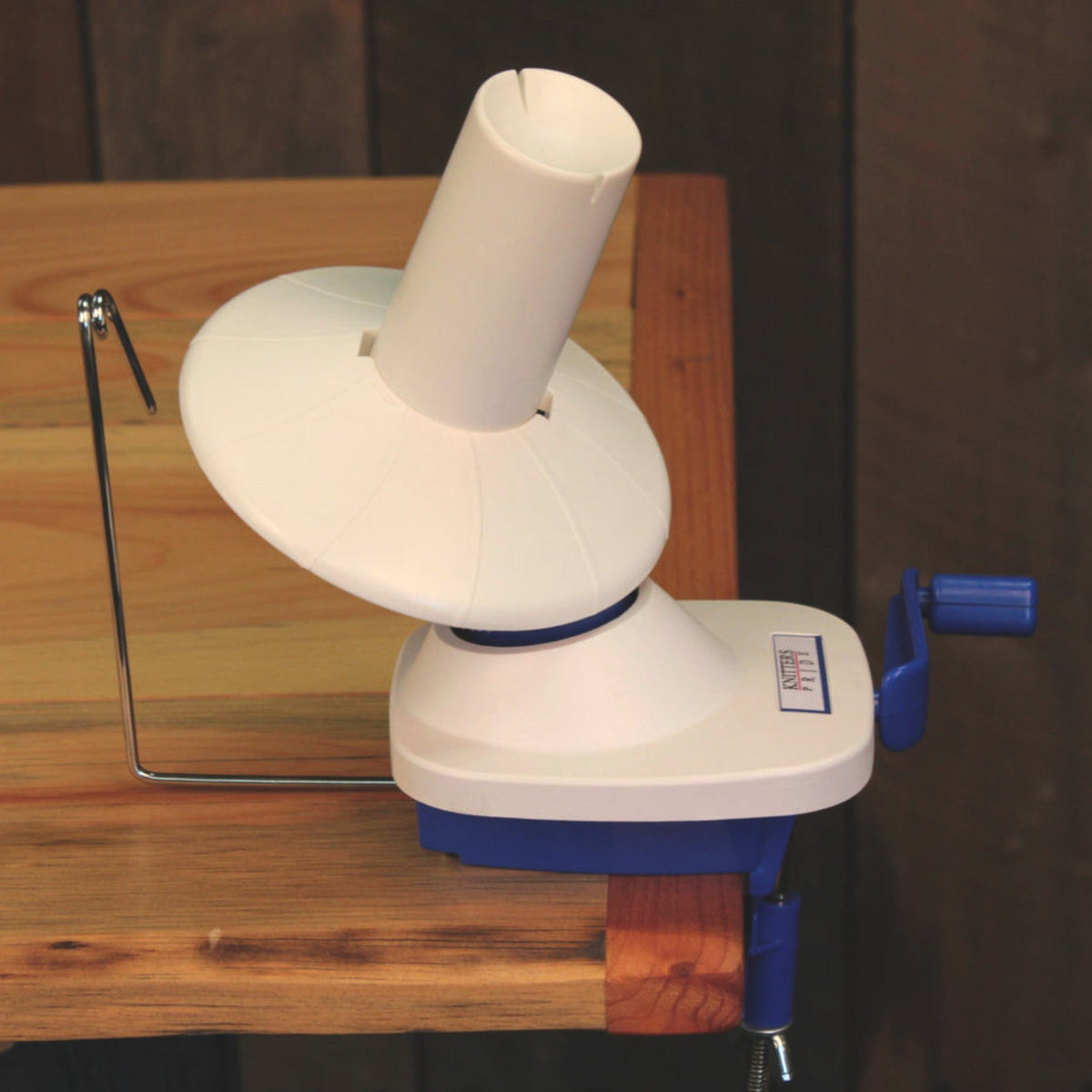 A white and blue Accessories Unlimited Knit Pro (Knitters Pride) Ball/Wool Winder is clamped to a wooden table, featuring a metal yarn guide and rotating spindle for winding yarn into neat balls.