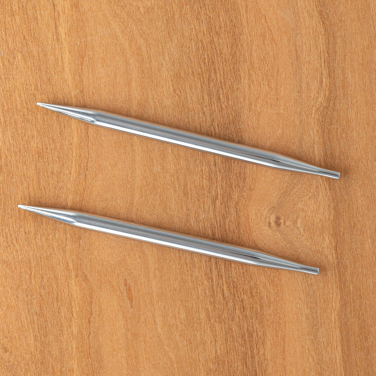 Two 5" Knit Pro (Knitters Pride) Nova Metal Interchangeable Knitting Needles by Accessories Unlimited lie parallel, pointed at both ends and positioned horizontally on a wooden surface.