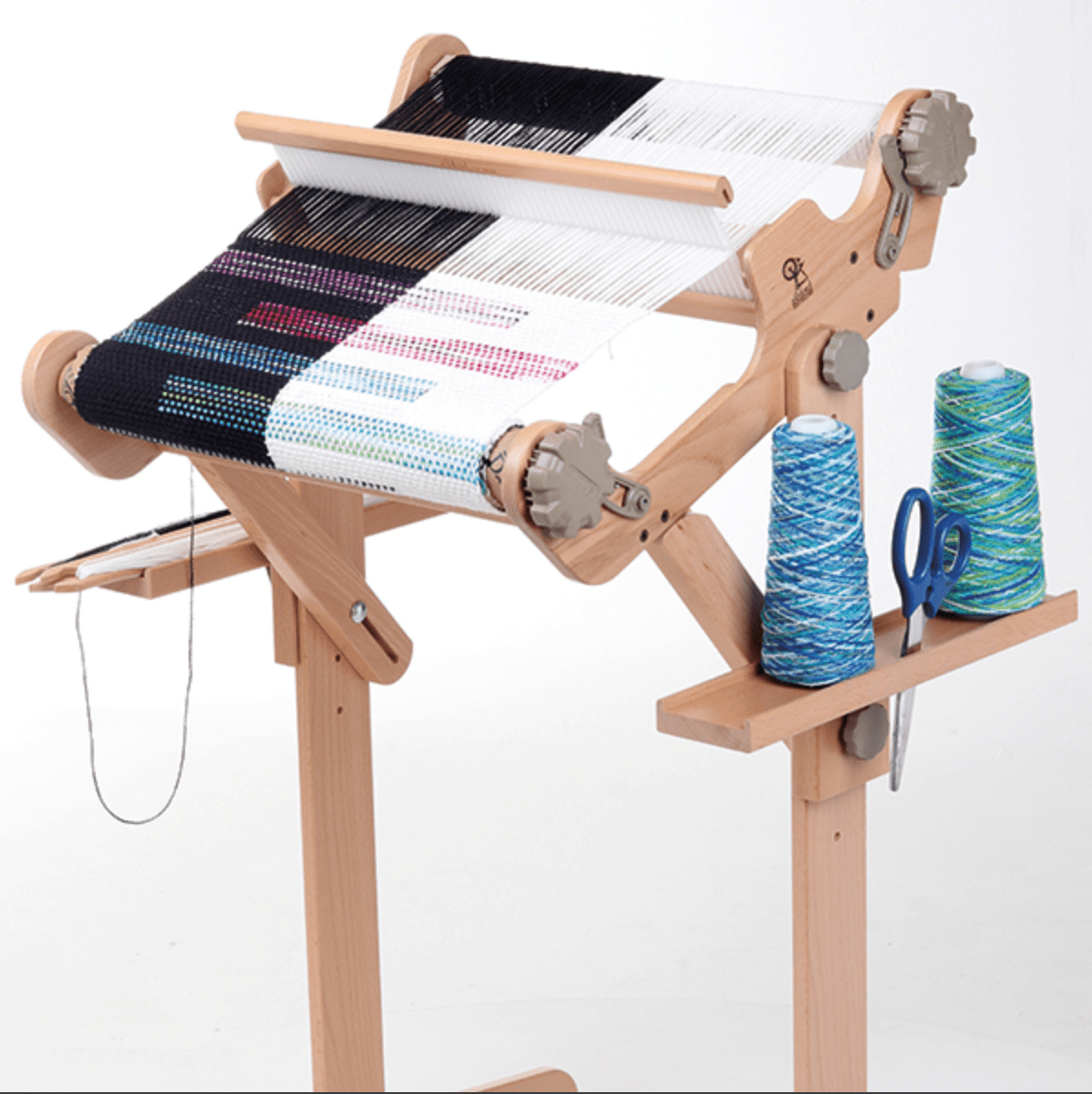A small wooden loom holds black, white, and colored threads in progress. Two blue and green yarn spools, scissors, and a weaving shuttle are organized in Ashford Rigid Heddle Loom Stand Accessories Trays by Ashford Handicrafts Limited.