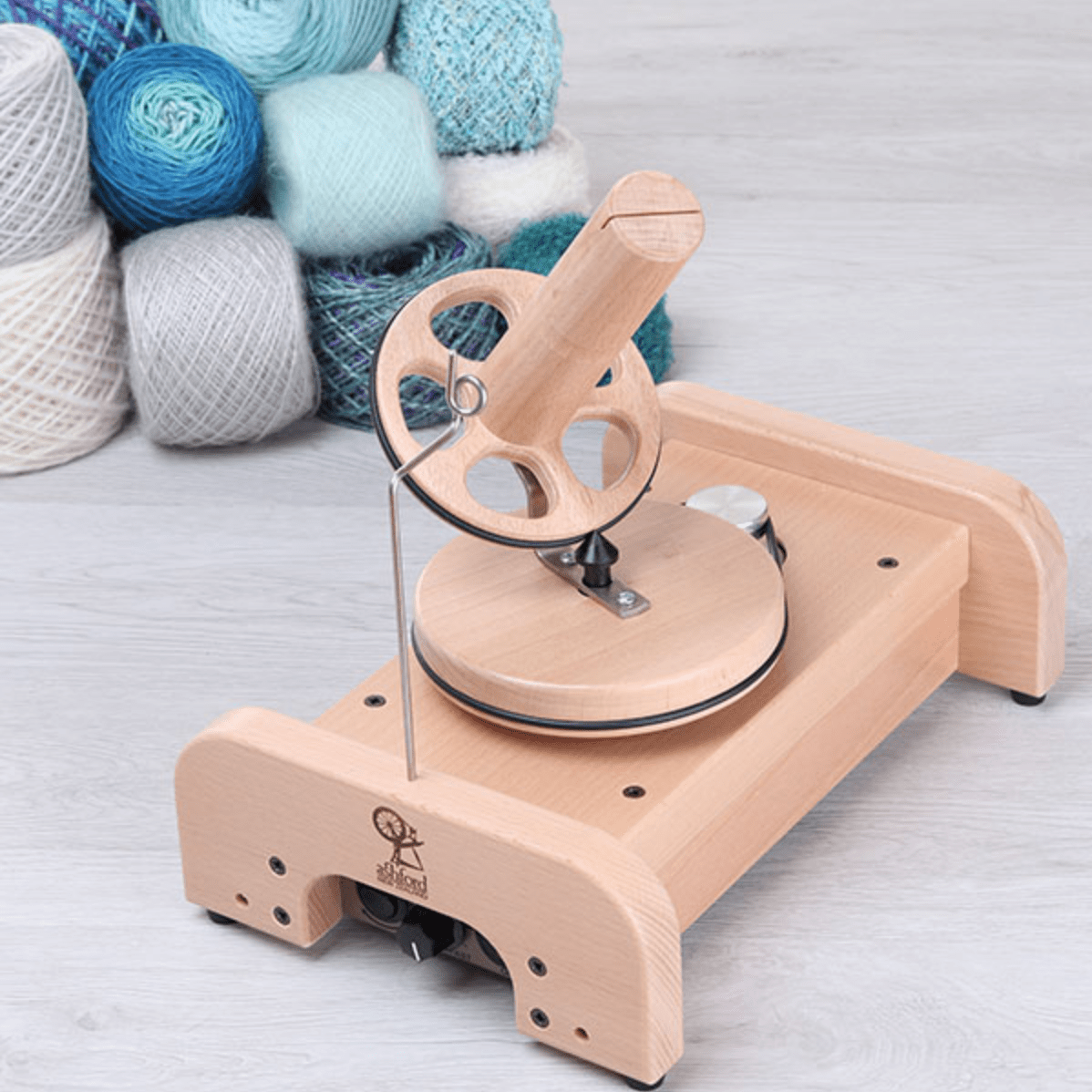 An Ashford Wooden Ball Winder by Ashford Handicrafts Limited rests on a light floor, surrounded by neatly stacked balls of blue, grey, and white yarn.