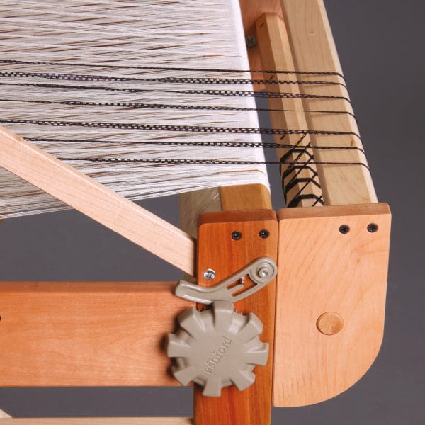 Close-up of the Ashford Second Back Beam for Table Looms by Ashford Handicrafts Limited, shown with white and black threads stretched across the loom. A gray plastic knob is attached to the side, and the frame displays both light and medium wood tones. The second back beam is partially visible behind the main weaving area, emphasizing the loom’s sturdy construction.