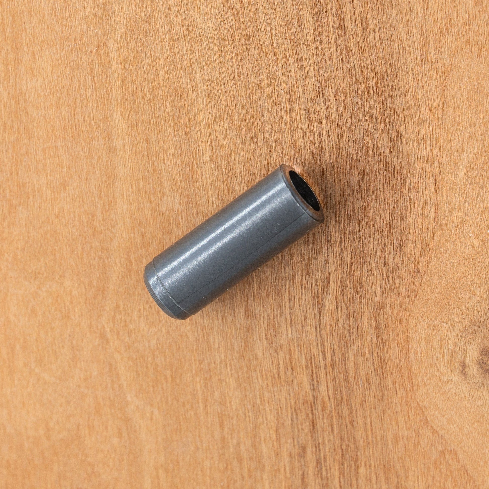 A small, cylindrical gray plastic tube of Ashford Maiden Bearing Replacement by Ashford Handicrafts Limited lies diagonally on a light brown wooden surface, ready for use with your Ashford Spinning Wheel.