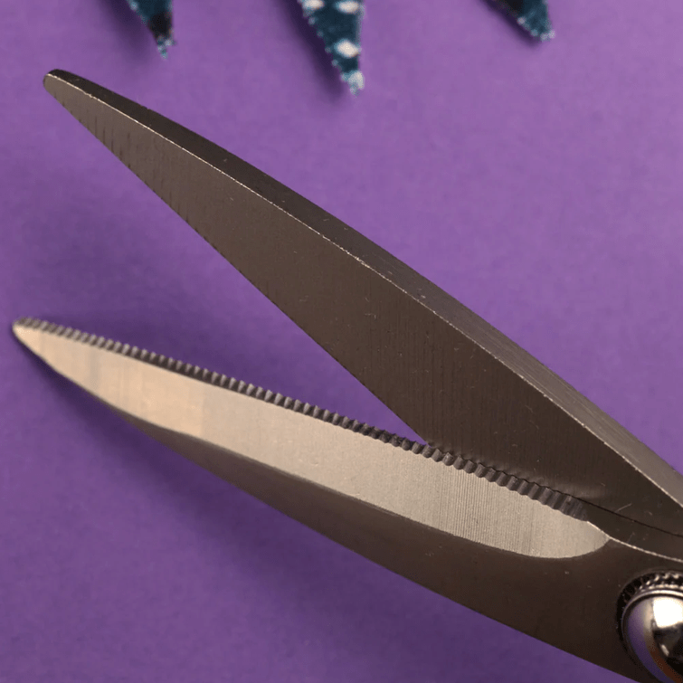 Close-up of Bryson Distributing, Inc. 6.75" Micro-Serated High-Precision Scissors with zigzag blades on a purple background, a small fabric swatch at the top displays the scissors' precise zigzag cut.