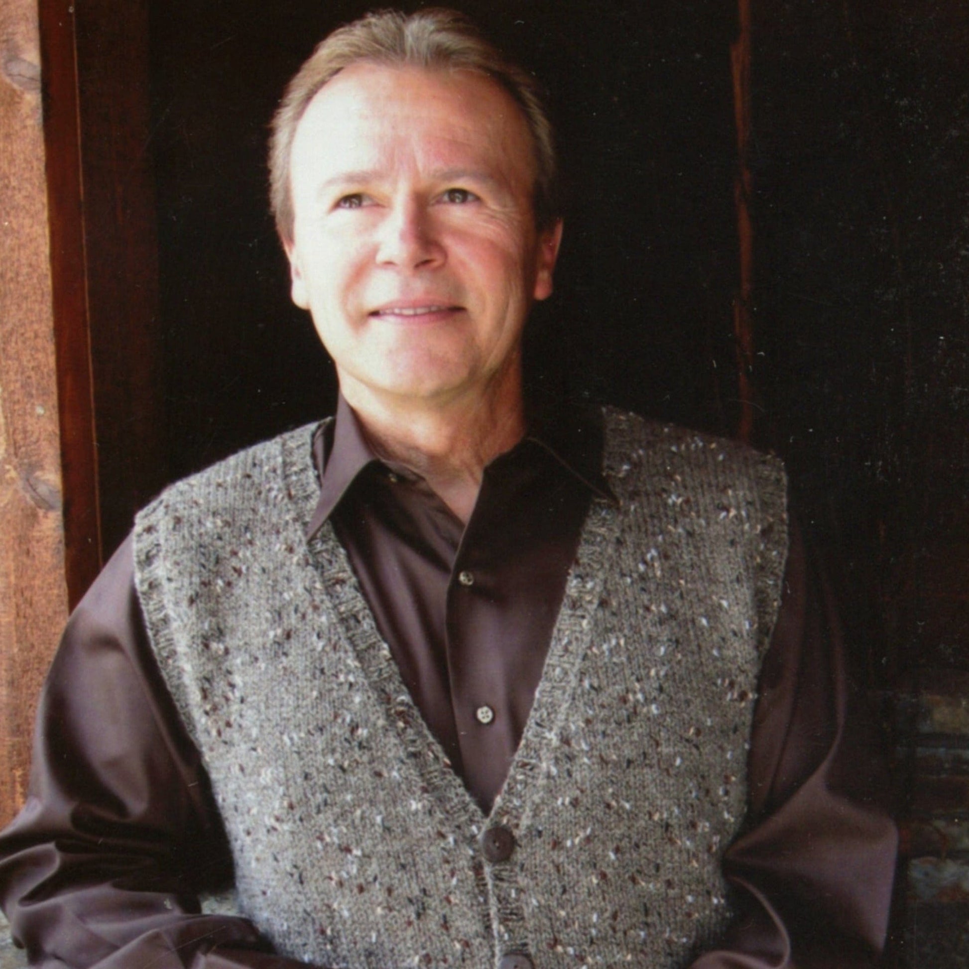 A man with light brown hair stands in a doorway, smiling slightly upward while wearing the Knitting Pure & Simple Basic Cardigan Vest for Men #276 (print) over a dark brown button-up shirt.