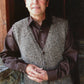 A man with short light hair, wearing a brown shirt and the Basic Cardigan Vest for Men #276, print by Knitting Pure & Simple, stands indoors near a stone wall and wooden doorway, looking up and smiling with hands clasped.