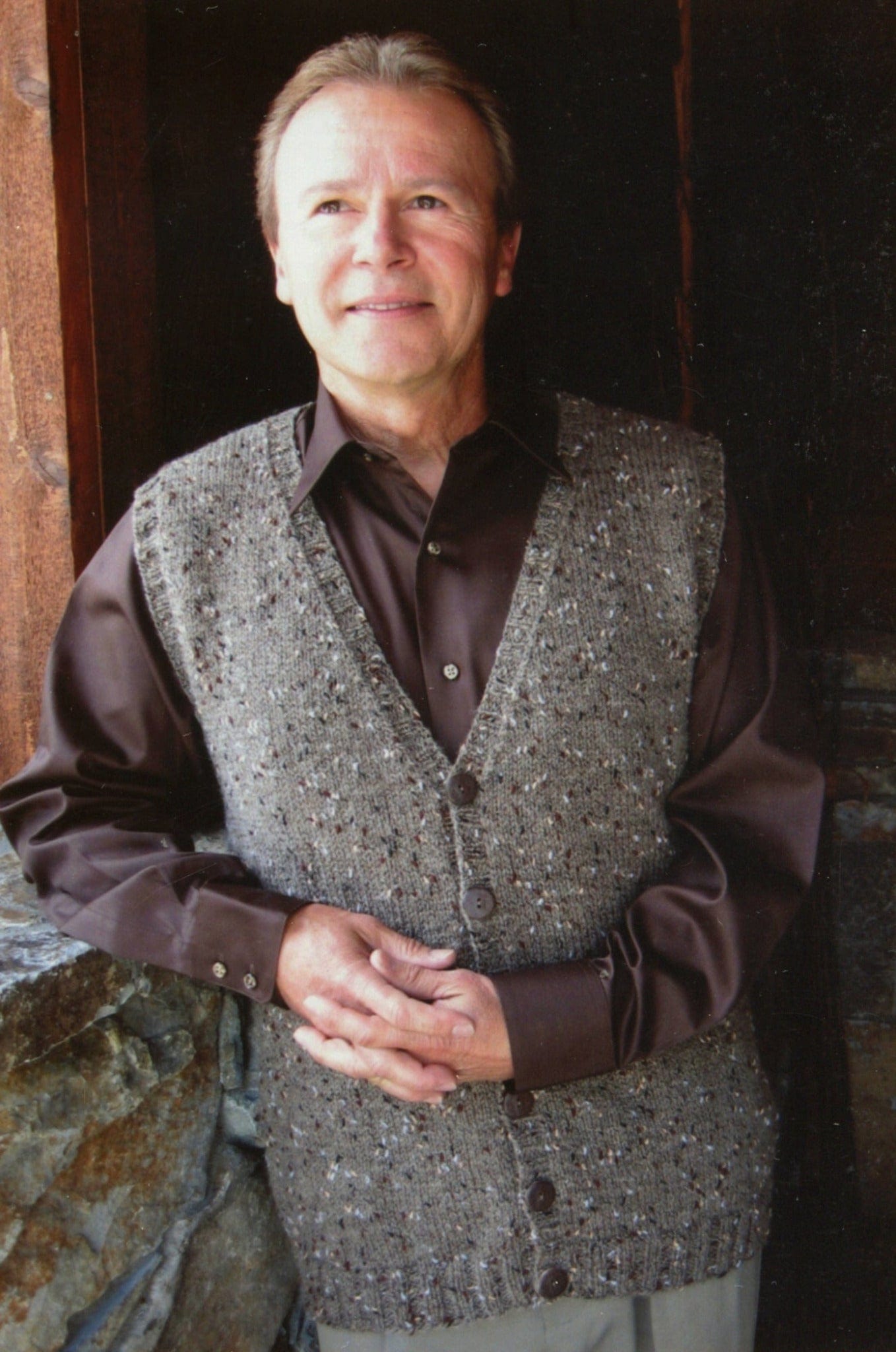 A man with short light hair, wearing a brown shirt and the Basic Cardigan Vest for Men #276, print by Knitting Pure & Simple, stands indoors near a stone wall and wooden doorway, looking up and smiling with hands clasped.