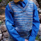 A child models the Basic Vest for Children, #256 by Bryson Distributing, Inc.—a blue and multicolored worsted weight sweater vest—over a blue button-up shirt outdoors near rocks, with only the torso and lower face shown.