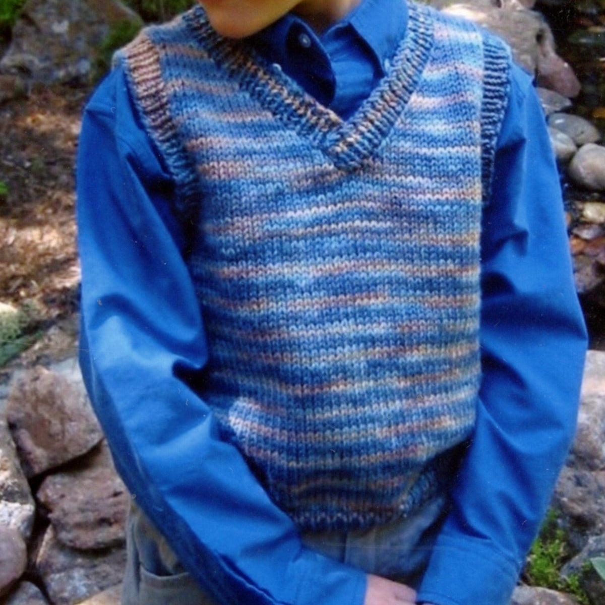 A child models the Basic Vest for Children, #256 by Bryson Distributing, Inc.—a blue and multicolored worsted weight sweater vest—over a blue button-up shirt outdoors near rocks, with only the torso and lower face shown.