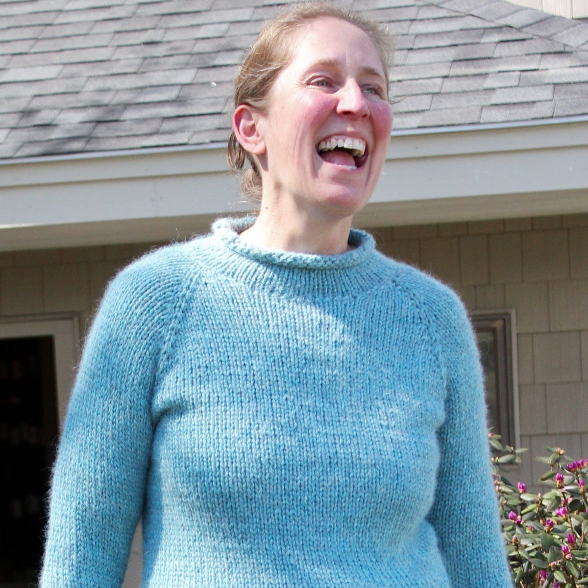 A woman wearing the Knitting Pure & Simple Bulky Neck Down Pullover #992 in light blue smiles and laughs outdoors near a building with a gray roof and beige siding, with some plants visible in the foreground.