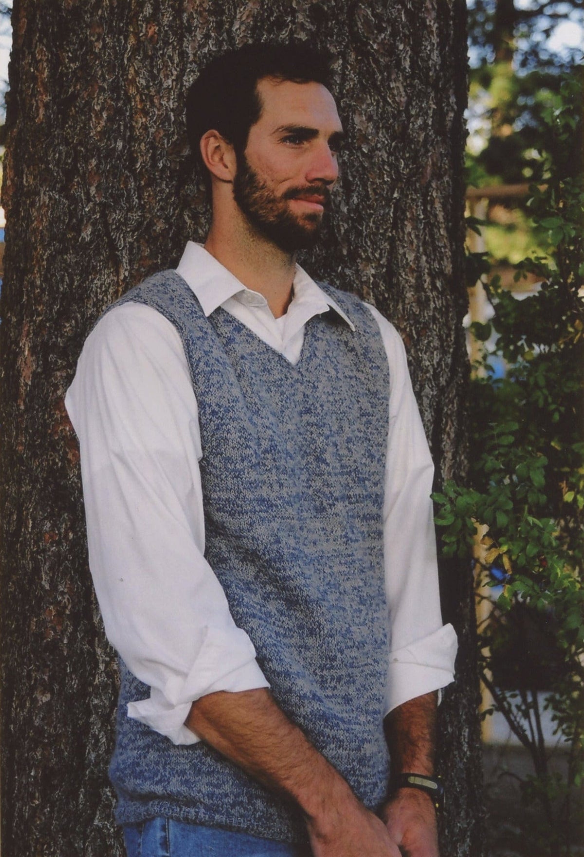 A man with a beard and short dark hair stands by a tree, wearing a white collared shirt and the Basic Vest for Men from Bryson Distributing, Inc. His sleeves are rolled up, and he gazes thoughtfully to the side.