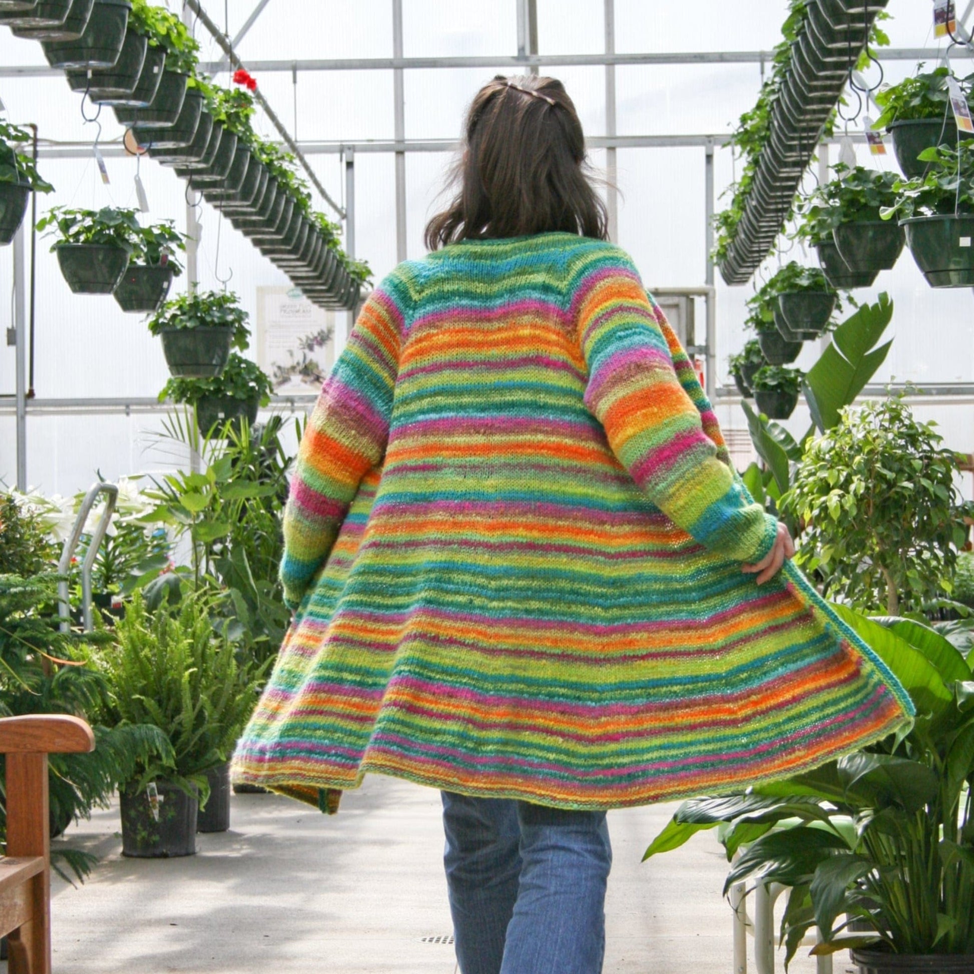 A person wearing the Neck Down Swing Coat (#285) by Bryson Distributing, Inc.—a long, colorful cardigan made from worsted weight yarn—walks through a greenhouse filled with lush plants and hanging pots, facing away from the camera.