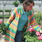 Wearing the Bryson Distributing, Inc. Neck Down Swing Coat (#285), a woman in a colorful, striped cardigan and blue shirt admires pink flowers among lush greenery in a greenhouse.