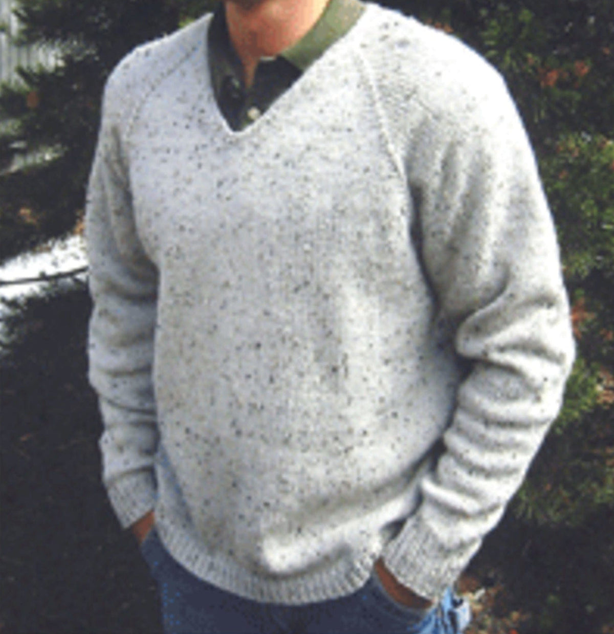 A person stands outdoors, hands in pockets, wearing the Bryson Distributing V-Neck Down Pullover for Men (#247) over a green collared shirt. Their face is partly out of frame, with trees and greenery visible in the background.