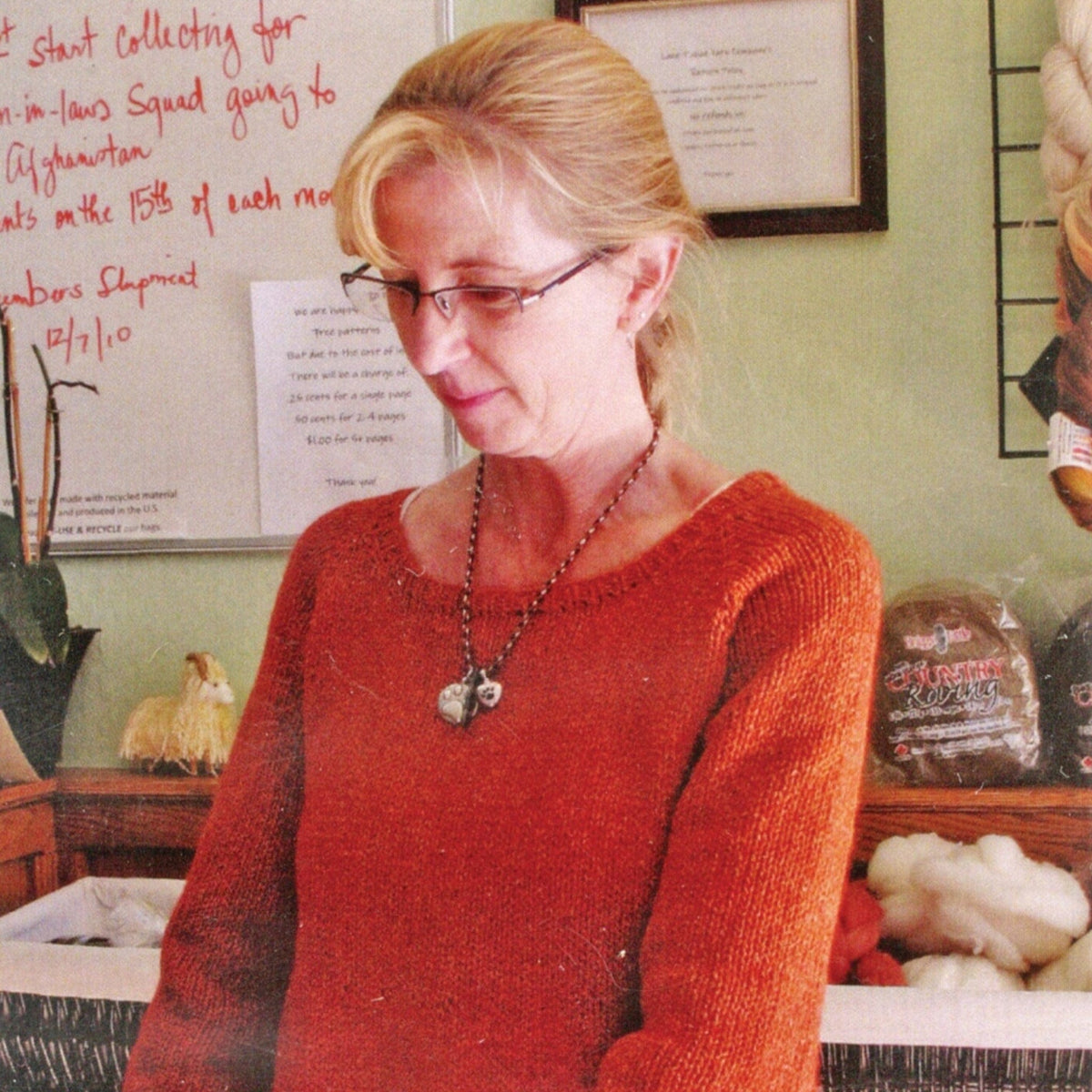 A woman with blonde hair in a ponytail, wearing glasses and a red-orange Top Down Swing Pullover #115 by Knitting Pure & Simple, stands indoors near a counter with handwritten notes and wool products visible in the background.