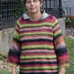 An older woman with short gray hair and glasses models the Top Down Swing Pullover, #115 by Bryson Distributing, Inc.—a colorful striped flared knit in worsted weight yarn—paired with blue jeans outdoors before a black iron fence and greenery.