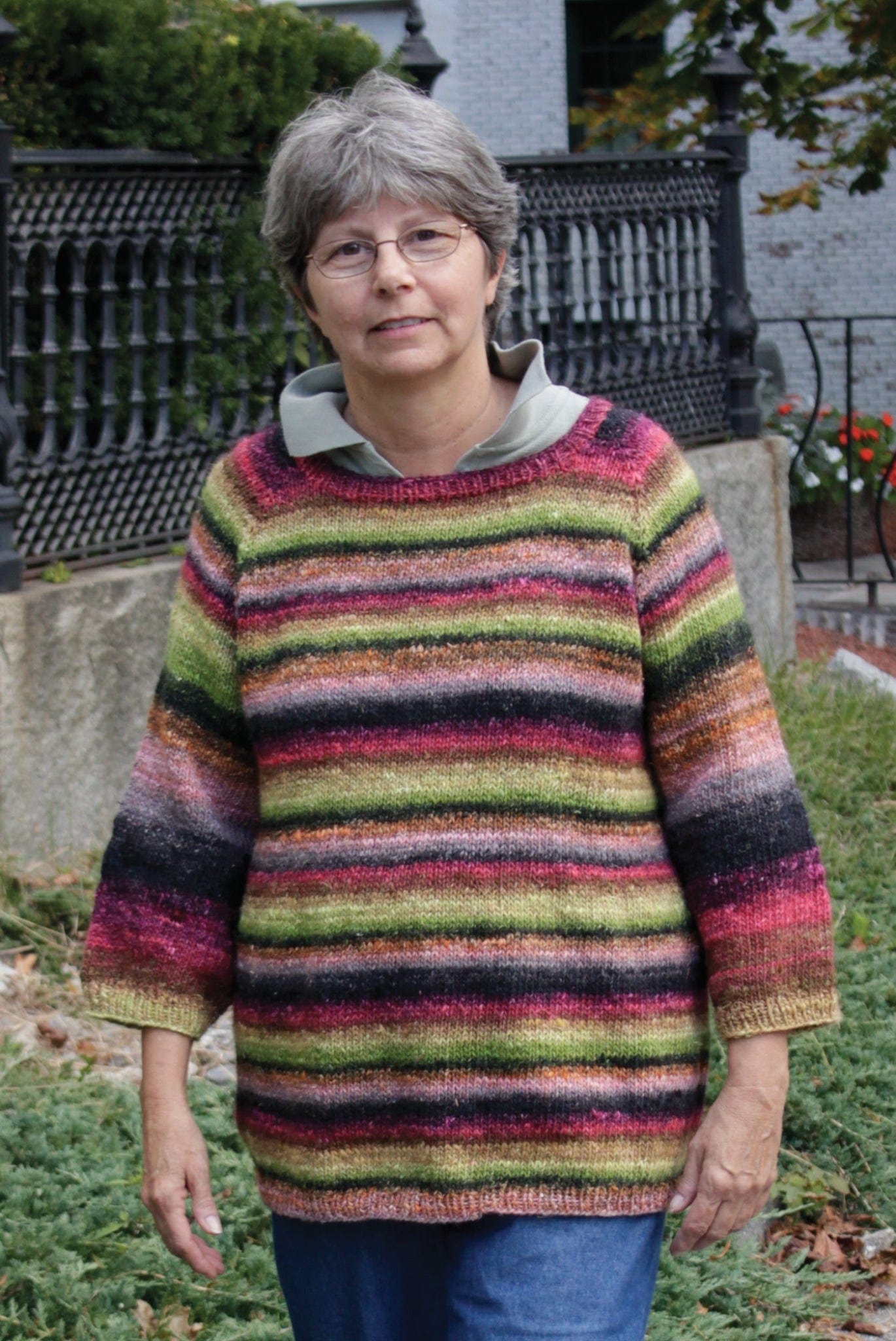 An older woman with short gray hair and glasses models the Top Down Swing Pullover, #115 by Bryson Distributing, Inc.—a colorful striped flared knit in worsted weight yarn—paired with blue jeans outdoors before a black iron fence and greenery.