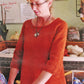 A woman in glasses and a Top Down Swing Pullover, #115 by Bryson Distributing, Inc., stands before shelves of worsted weight yarn and a handwritten notice, looking down while holding something out of view.