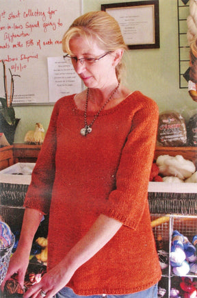 A woman in glasses and a Top Down Swing Pullover, #115 by Bryson Distributing, Inc., stands before shelves of worsted weight yarn and a handwritten notice, looking down while holding something out of view.