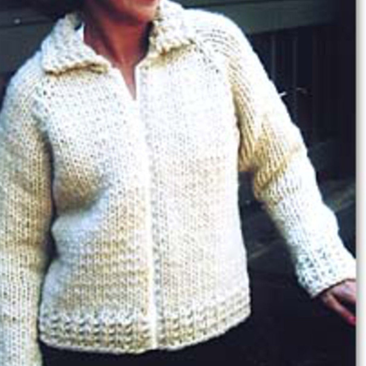 A person wears the Weekend Neck Down Jacket #234 by Bryson Distributing, Inc.—a thick, off-white, hand-knit cardigan with a collared neck, textured pattern, front opening, and long sleeves—while standing outdoors.