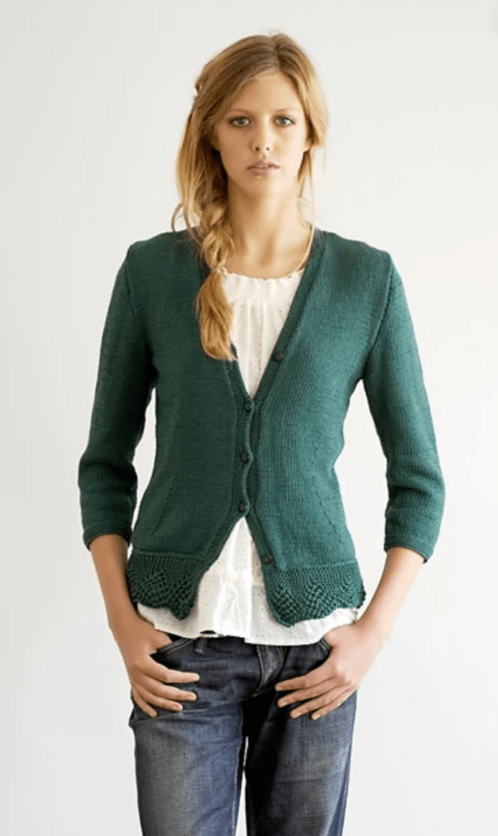 A woman stands against a white background, wearing the Fitted Cardigan by Jo Sharp (print only) from Yankee Knitter Designs over a white blouse and blue jeans, her hands on her hips with a neutral expression.