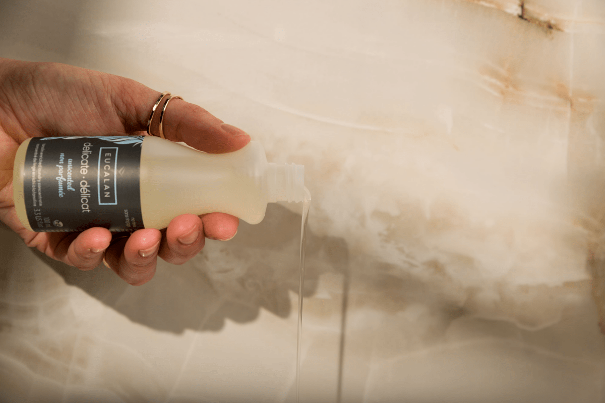 A hand adorned with rings holds a 100 ml (3.3 oz) Eucalan Wool Wash bottle and pours clear liquid onto a marble surface. This no-rinse, lanolin-enriched concentrate from Eucalan is a biodegradable textile cleanser, perfect for preserving your most cherished fabrics.