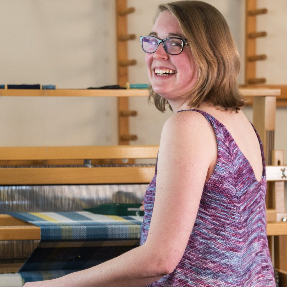 Wearing the Flower In The Sun Tank Top by Sierra Roberts from Halcyon Yarn, a woman smiles as she weaves a colorful textile at a loom. The downloadable knit top is perfect for creative days at home.