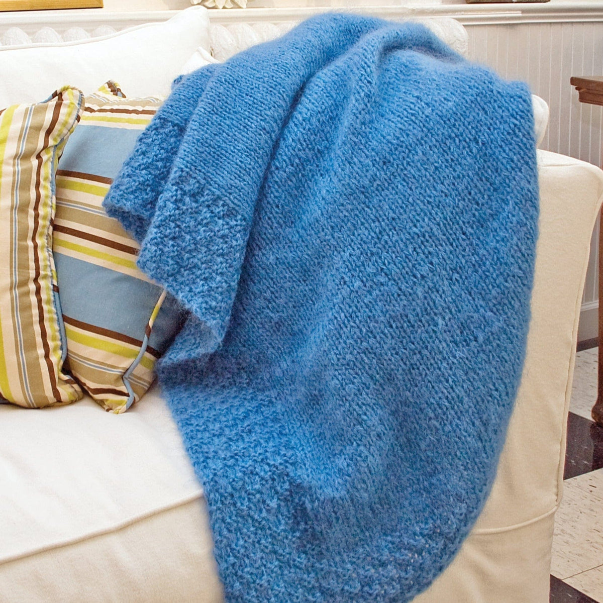 A Victorian Luxury Throw in a fluffy blue brushed mohair by Halcyon Yarn is draped over a cream-colored sofa, paired with striped and patterned throw pillows.