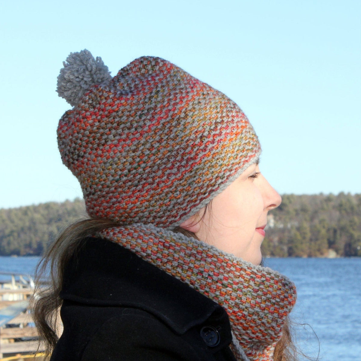 A woman in the Telephone Line Hat & Cowl by Halcyon Yarn, knit in a textured slip stitch, stands outdoors by a lake. She wears a multicolored set with pom-pom and scarf, gazing sideways in her dark coat with trees in the background.
