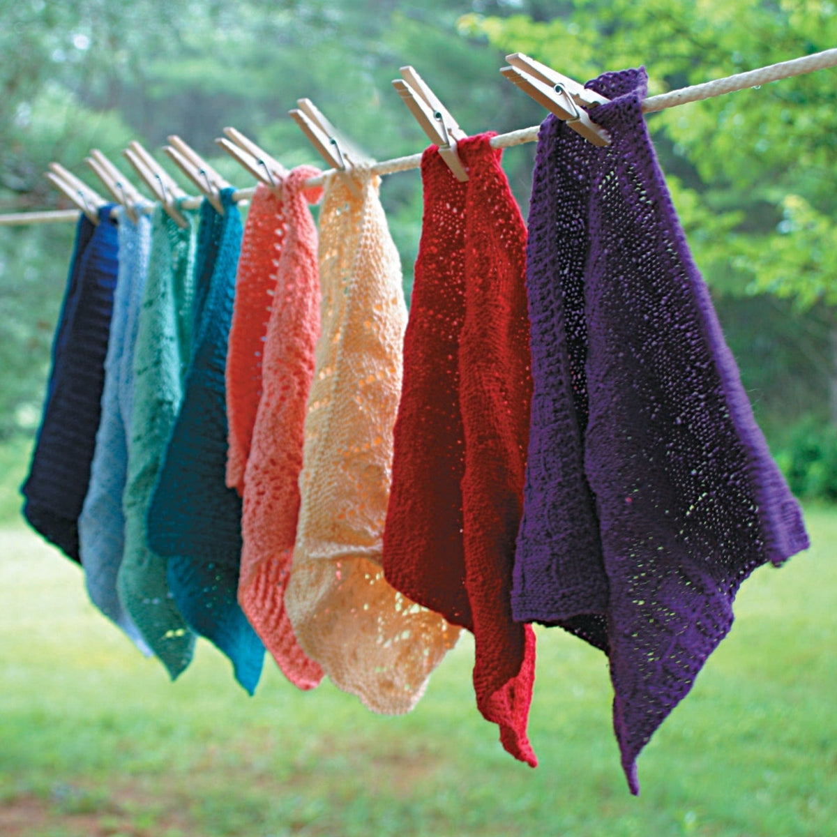 Seven colorful sport weight cotton washcloths, made with Halcyon Yarn Casco Bay, hang on an outdoor clothesline. Get the Casco Bay Washcloths downloadable pattern from Halcyon Yarn—perfect for testing new wash cloth designs!.