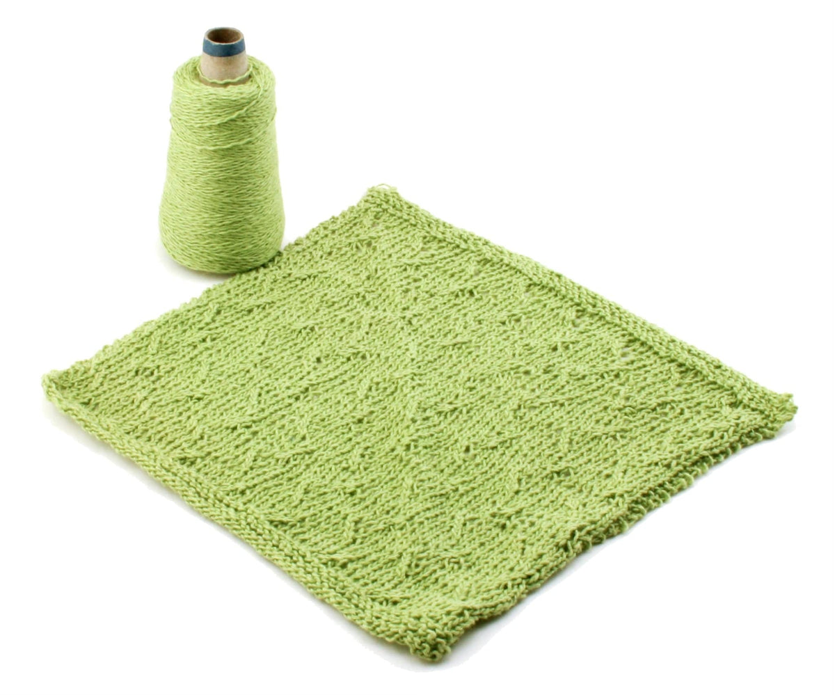 A Casco Bay Washcloths download by Halcyon Yarn is shown with a green knitted washcloth featuring a subtle wave pattern, alongside a cone of sport weight cotton yarn, all displayed on a white background.