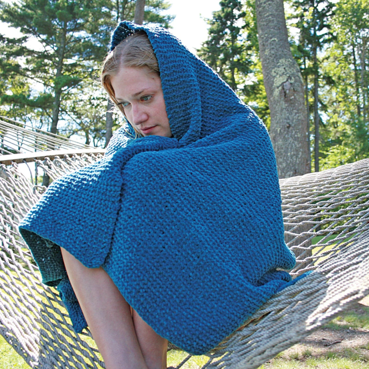 A person sits curled up on a hammock outdoors, wrapped in the Halcyon Yarn Casco Cottage Chenille Throw. With its plush hooded design and machine-washable fabric, it offers cozy comfort amidst trees and sunlight in a serene setting.