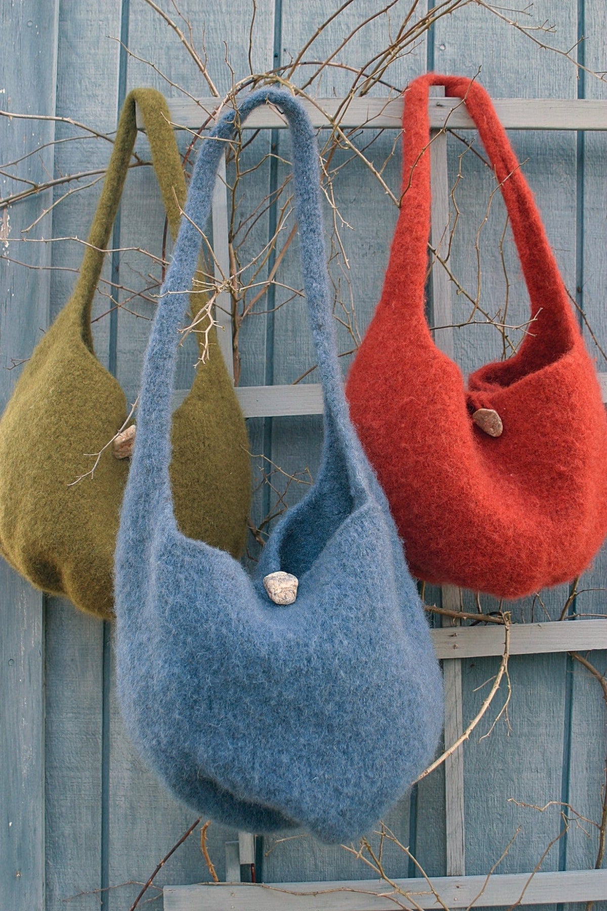 Three Halcyon Yarn Felted Satchel Knitted bags in green, blue, and red hang on a rustic blue fence. Each features bulky wool construction with wooden button closures, framed by bare twigs.