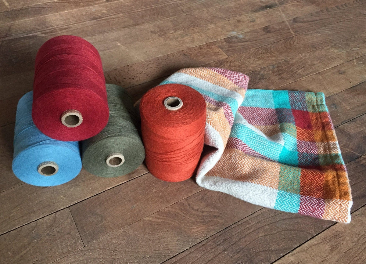 Four spools of yarn in red, blue, olive, and orange sit beside a vibrant geometric handwoven towel from Halcyon Yarn’s Eight Organic Cottolin Towels collection, all arranged on a wooden floor.