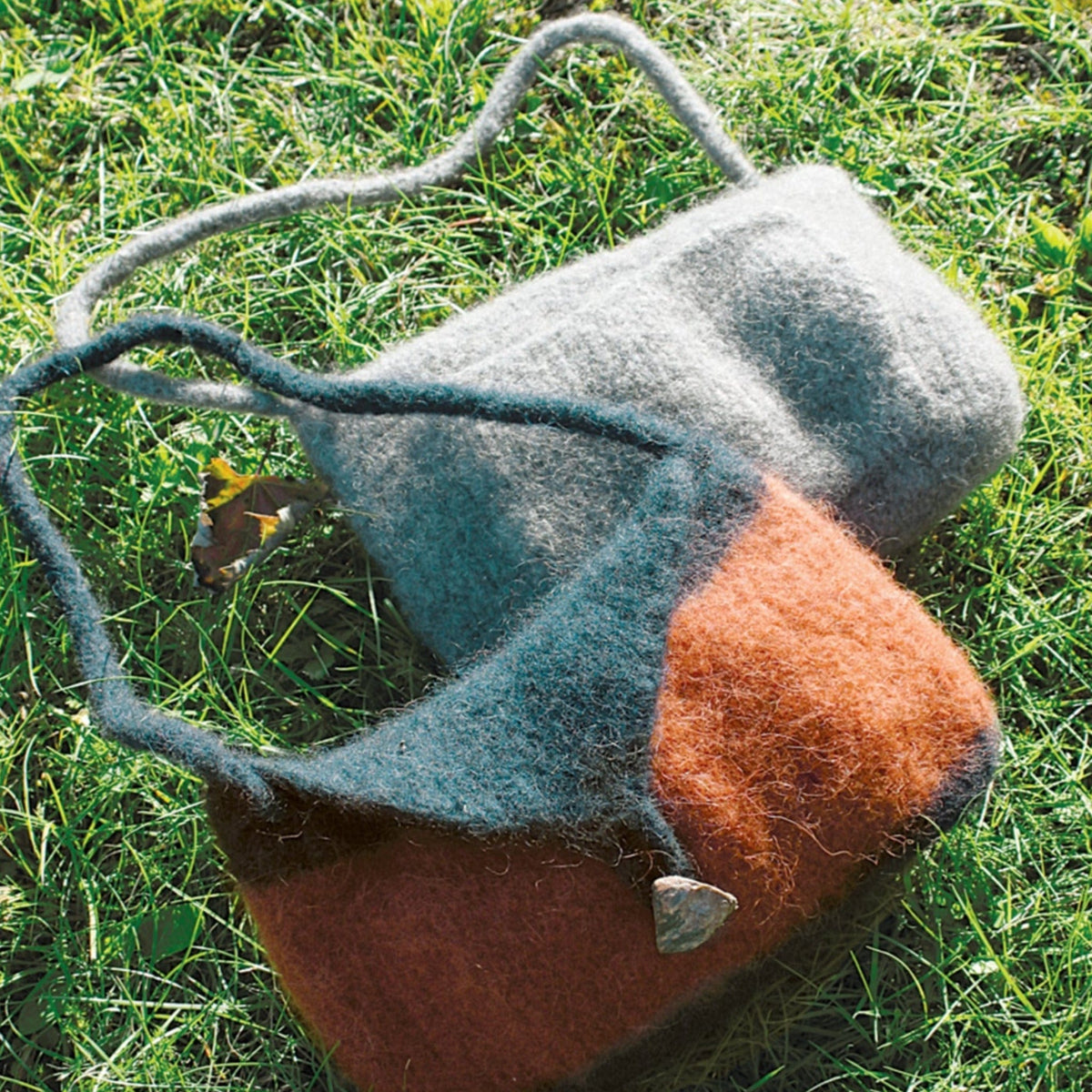 Two Halcyon Yarn Knitted Felt Skinny Strap Shoulder Bags rest on green grass: one is solid gray, the other rust and gray with a stone-shaped button. Both feature thick, soft straps—great inspiration for your next knitted felt purse project.