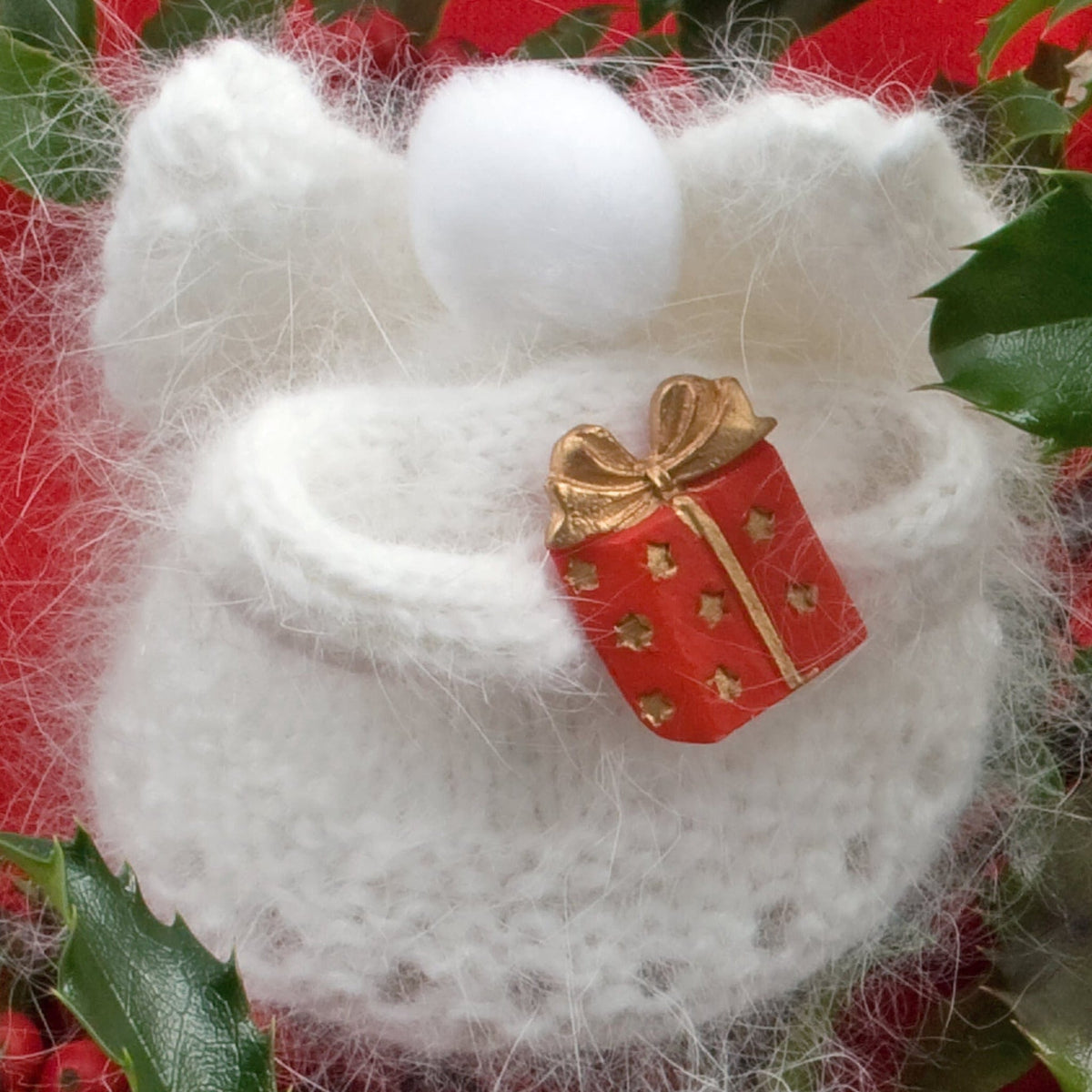 The Snow Angels Pattern by Halcyon Yarn exudes handmade elegance, showcasing a white knitted angel figure gently holding a small red gift adorned with a gold ribbon, nestled among vibrant poinsettia flowers and lush holly leaves, perfectly capturing the essence of timeless holiday decor.