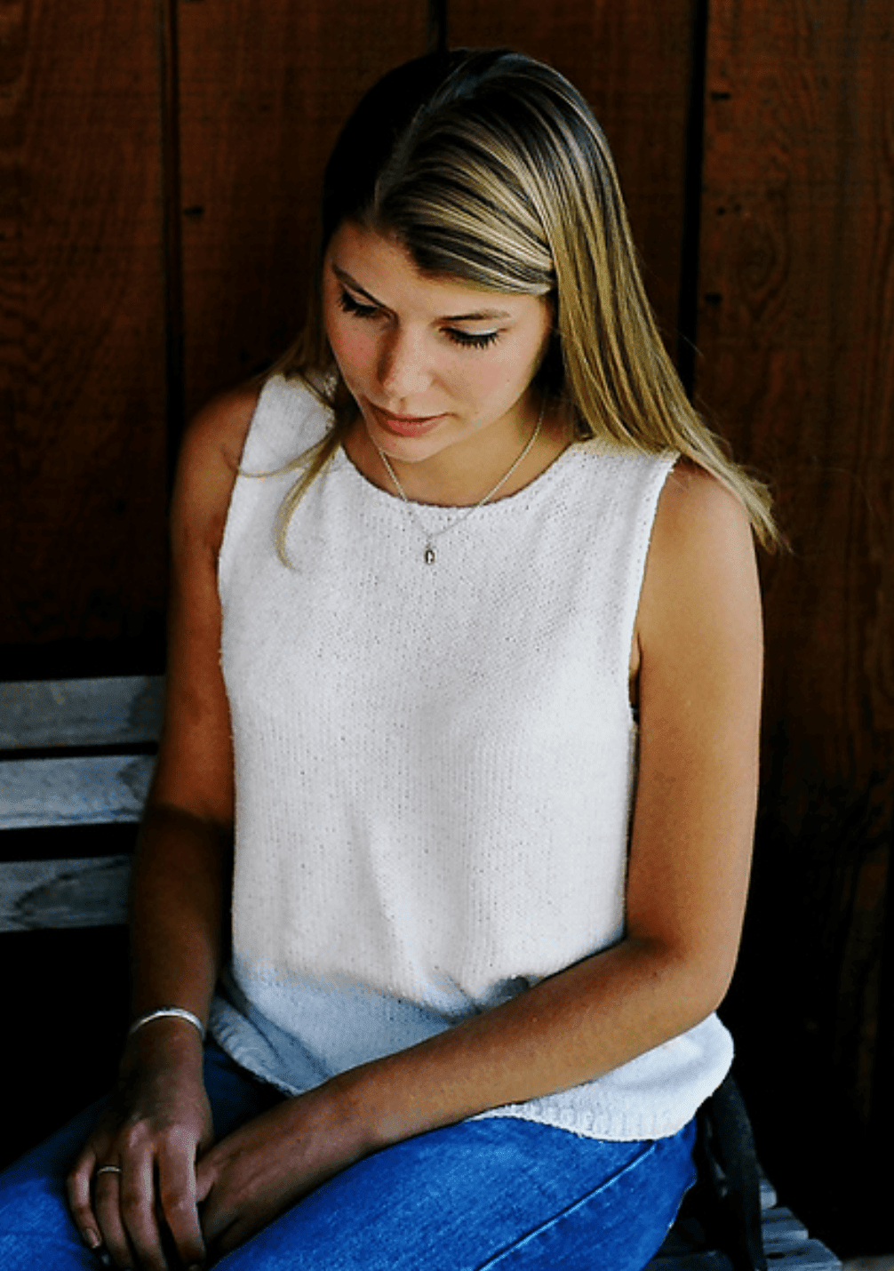 A young woman with long blonde hair sits on a bench, wearing the Basic Shell Tank Top Pattern (#997) by Knitting Pure & Simple and blue jeans, against a wooden wall background. She gazes downward thoughtfully.