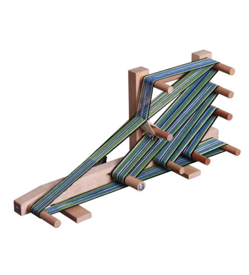 A wooden inkle loom with green, blue, and white striped threads tightly wound around its pegs, set on a plain white background. The loom, used for weaving narrow fabrics, showcases the intricate band of threads in progress.