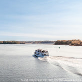 A small white ferry with passengers cruises Merrymeeting Bay on the Halcyon Yarn Cast Off & Cast On: 3-Hour Six Rivers Cruise (Sept. 13), gliding past trees under a clear blue sky.