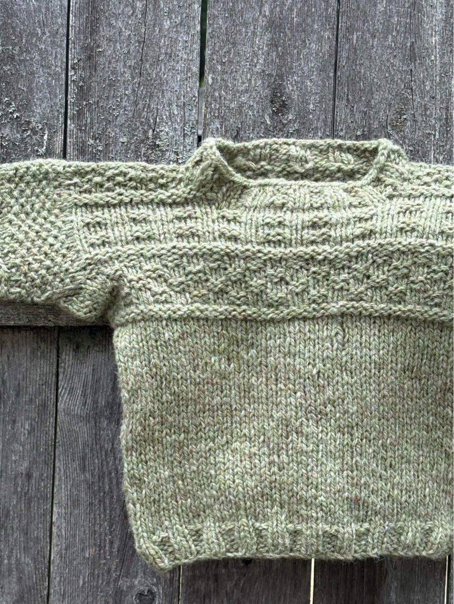 A mini sweater knit from Halcyon Yarn’s "Everything Gansey: Knit Your Own Mini Sweater with Linda Perry (October 3, 2026)" is laid flat on a weathered wooden surface, showcasing its light green color and textured gansey patterns on the sleeves and chest.