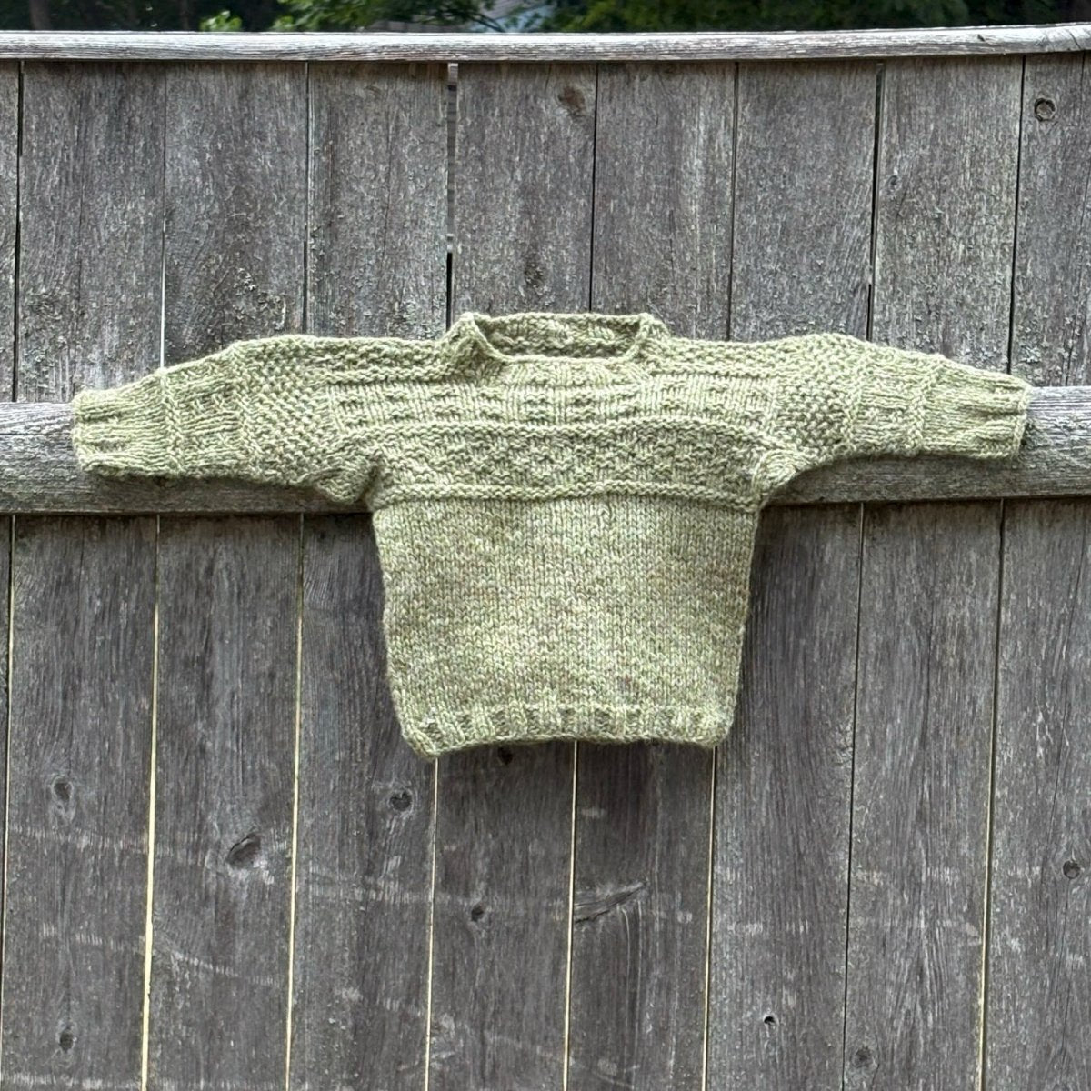 A hand-knitted olive green mini sweater, created using the "Everything Gansey: Knit Your Own Mini Sweater with Linda Perry (October 3, 2026)" kit from Halcyon Yarn, is displayed hanging on a rustic wooden fence. The sleeves are spread out to showcase the textured knit patterns inspired by traditional British ganseys on the upper part.