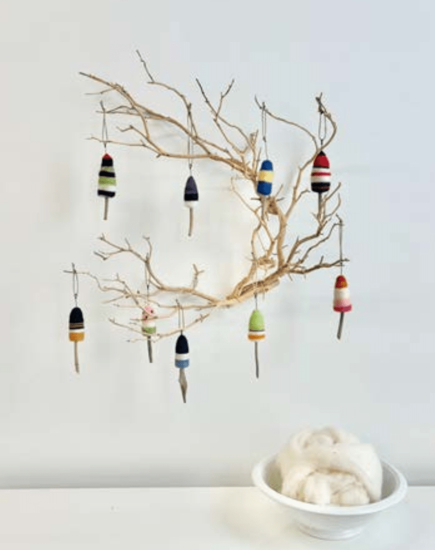 A minimalist scene features a bare branch on a white wall with colorful striped ornaments, including one inspired by the Driftwood Buoys Needle Felting Class with Michelle Provençal from Halcyon Yarn. Below, a bowl of wool evokes beginner felting materials.