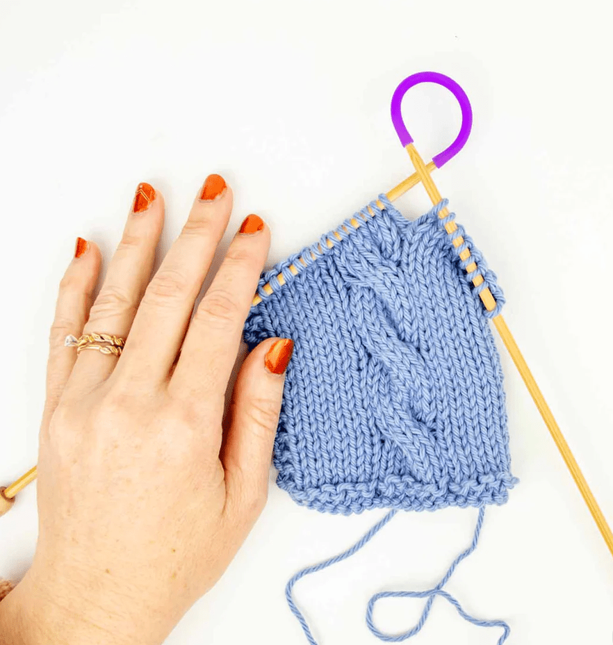 A hand with orange nails and gold rings rests near blue cabled knitting on wooden needles, secured with purple stitch holder and Twice Sheared Sheep Tip Ties - Needle Point Protectors covering the needle tips.