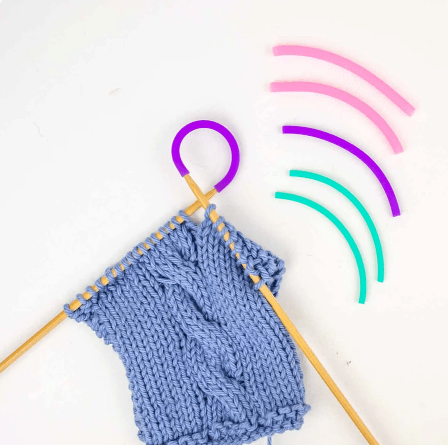 A blue knitted piece on wooden needles features Twice Sheared Sheep Tip Ties - Needle Point Protectors in purple, pink, magenta, and teal, along with colorful stitch holders and silicone tubes, all arranged on a white background.