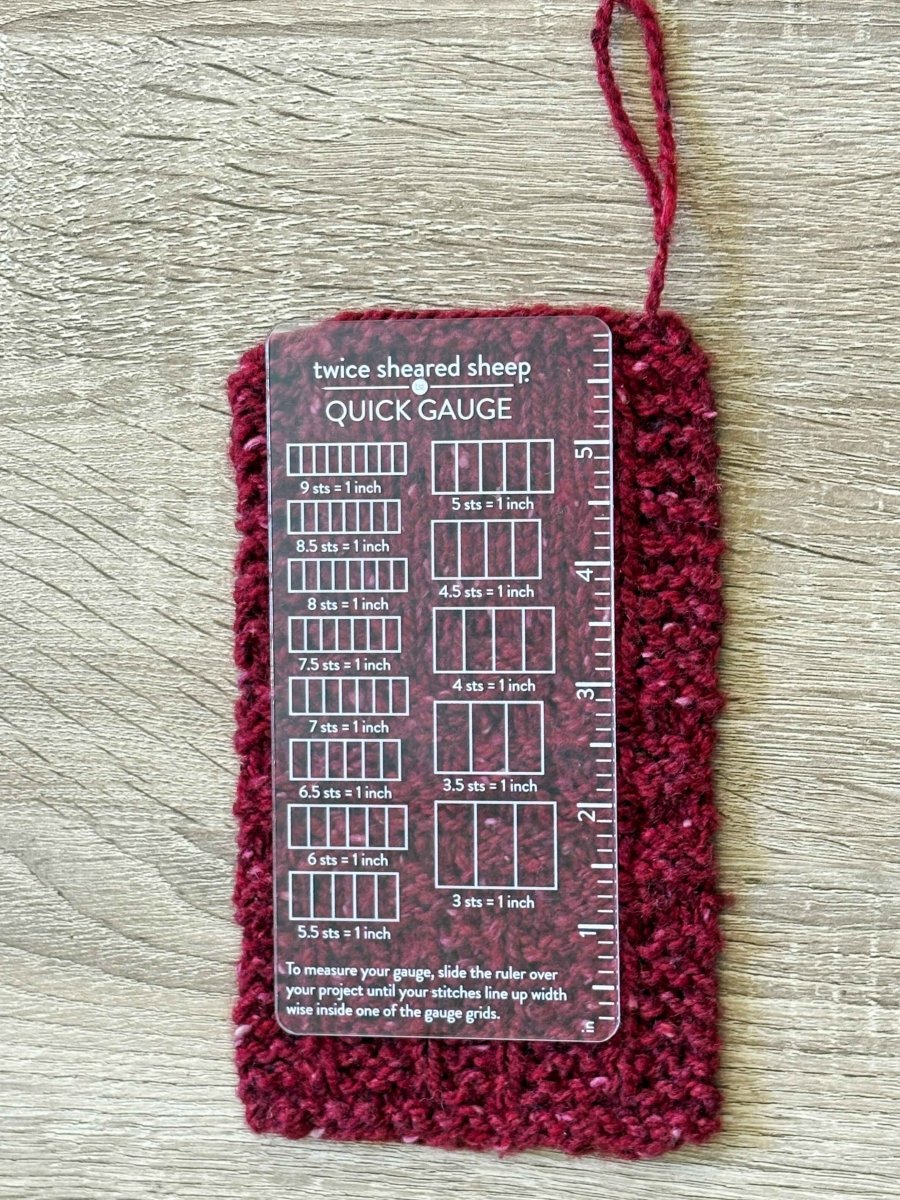 A Twice Sheared Sheep Quick Gauge Ruler, made of clear acrylic, rests on a maroon knit swatch to measure stitches per inch and row count using the ruler’s marked sections.