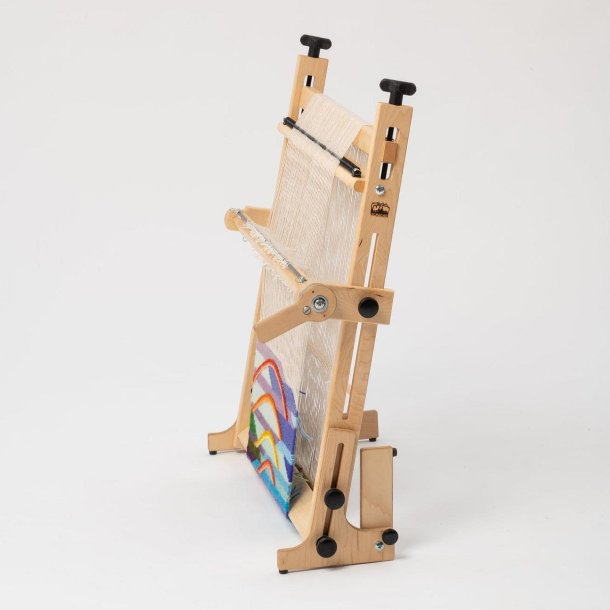 The Arras 20″ Tapestry Loom by Schacht Spindle Co. stands upright on a plain white background, displaying its wooden tabletop frame with adjustable gears for precise tension control. White warp threads are visible on the loom, which holds a partially woven, colorful fabric.