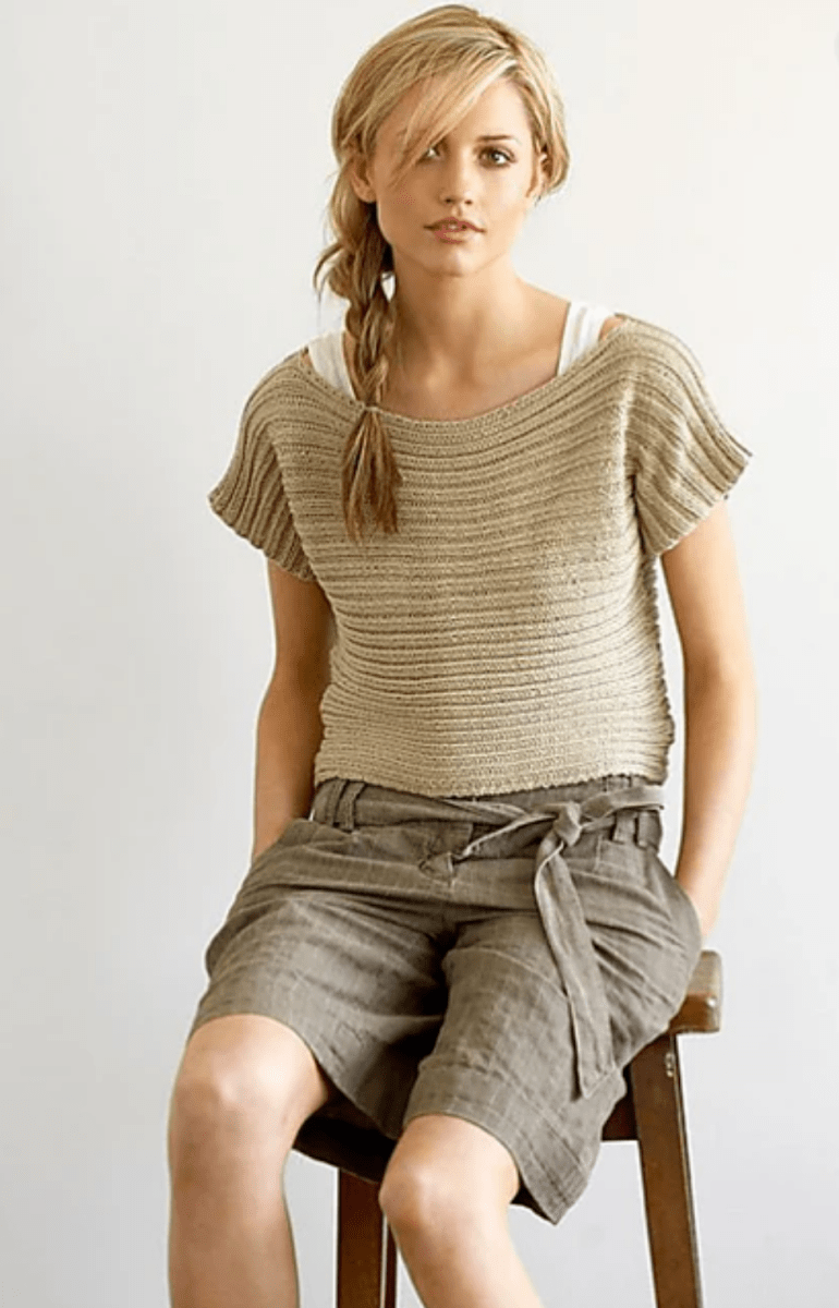 A young woman with blonde hair in a side braid sits on a wooden stool wearing the Sideways Ribbed Top Pattern by Jo Sharp, available in print only from Yankee Knitter Designs, layered over a white tank and tan high-waisted shorts. The background is plain and light.