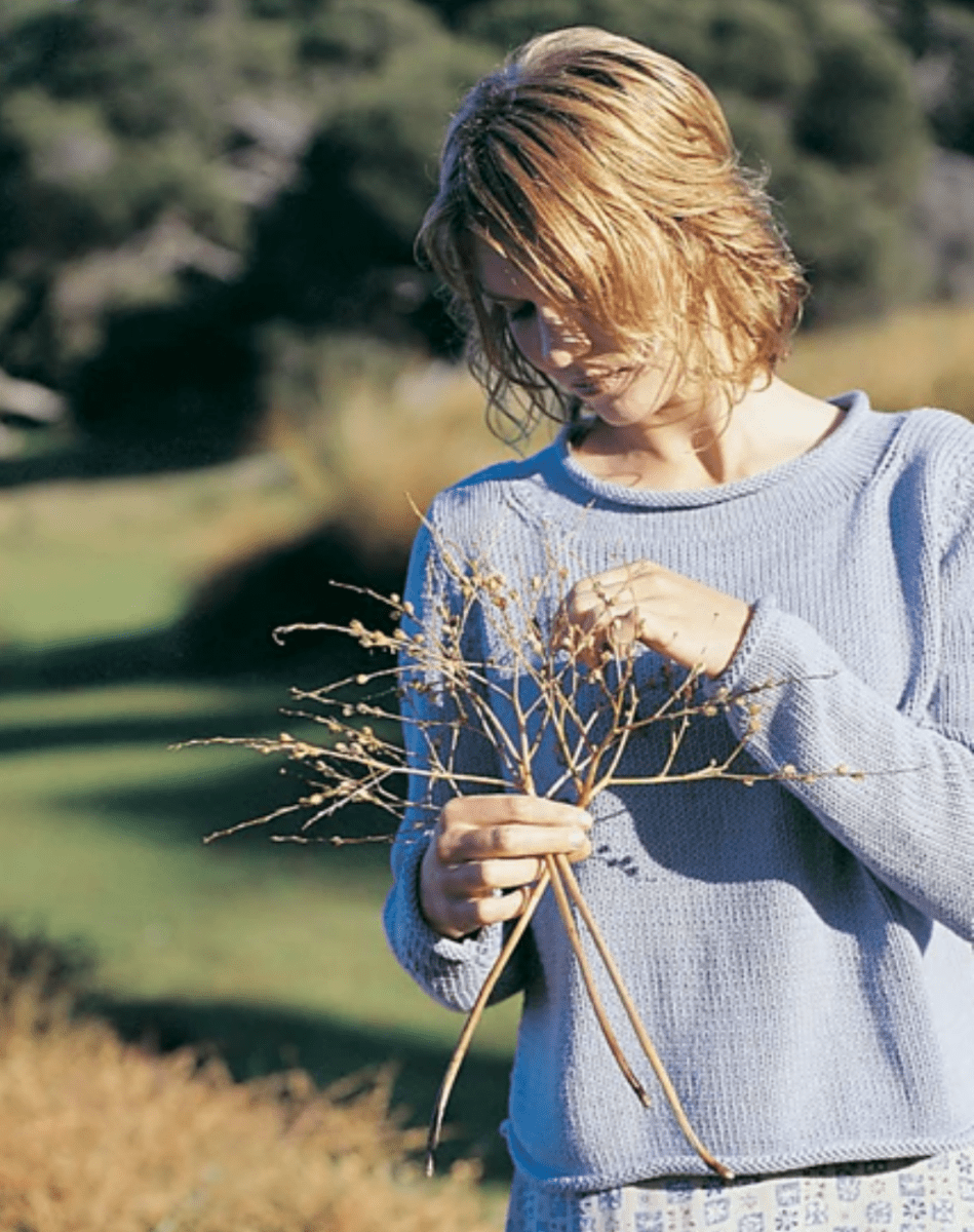 A person with short, light hair wears the relaxed-fit Somerville Sweater by Jo Sharp (print only) from Yankee Knitter Designs, standing outdoors in a sunlit grassy area and gently holding dried wildflowers.