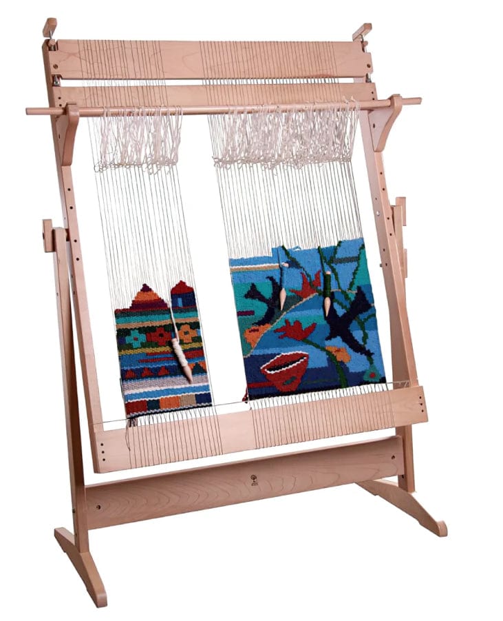 A wooden weaving loom displays a partially completed colorful tapestry. The tapestry features abstract and geometric designs in vibrant shades of blue, red, green, and orange. White warp threads are visible, with some areas of the design still unfinished.
