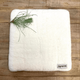 A square, white Woolbuddy Needle Felting Mat Large by Woolbuddy sits on a wooden surface with a small green pine branch at the top left and a "woodwoow" tag attached to the bottom right corner.