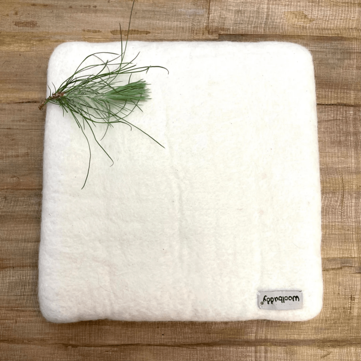 A square, white Woolbuddy Needle Felting Mat Large by Woolbuddy sits on a wooden surface with a small green pine branch at the top left and a "woodwoow" tag attached to the bottom right corner.